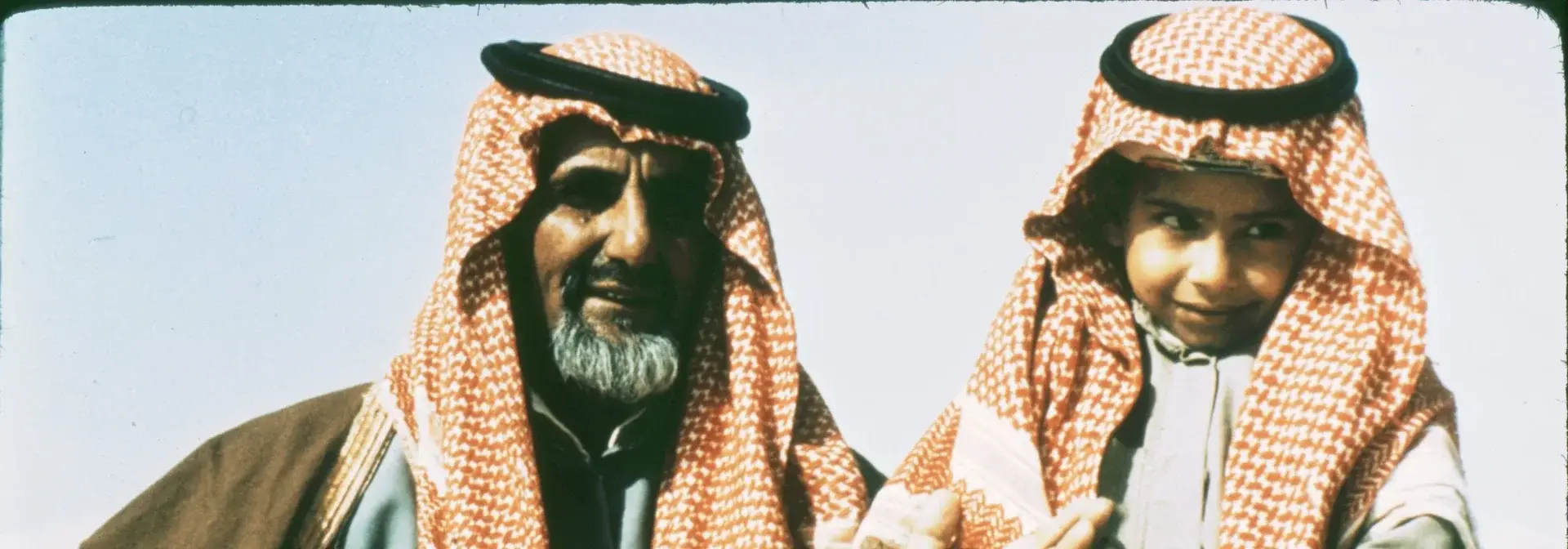 A Bedouin father and his son wearing Saudi traditional dress: a red-checked shemagh with a black agal, a thobe and a brown bisht.