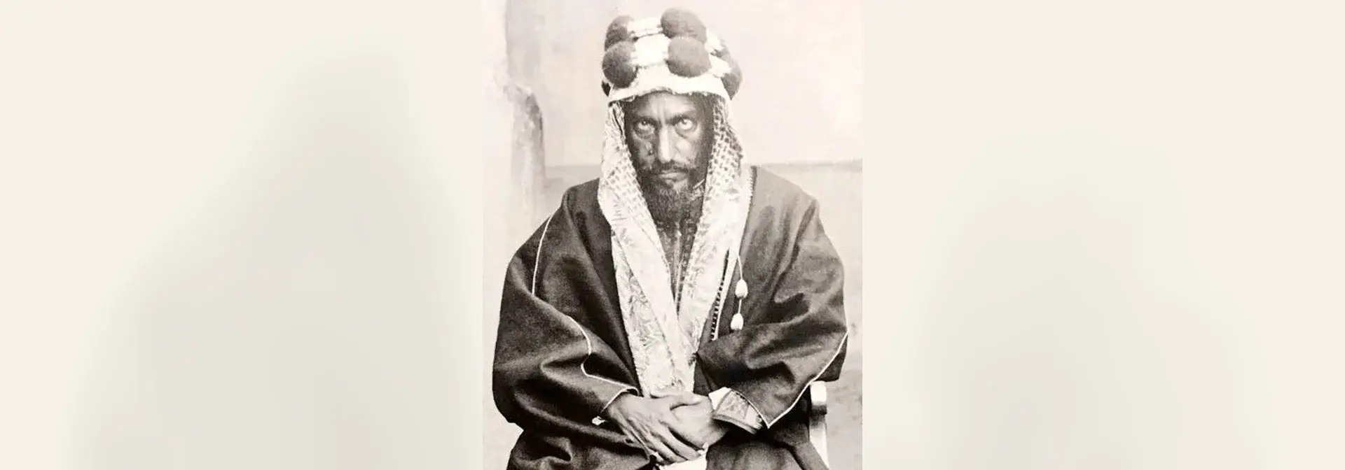 Historical portrait of Imam Abdulrahman bin Faisal Al Saud — a bearded elderly man wearing a ghutra with a pom-pom-trimmed agal and a dark gold-trimmed bisht, seated with his hands clasped