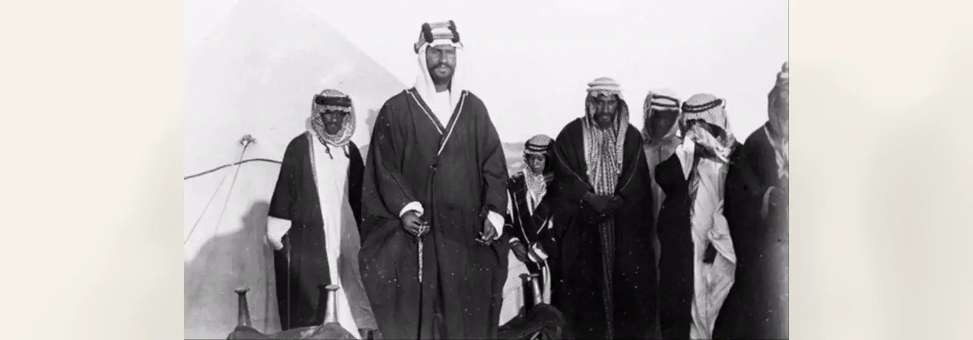 A 1915 historical photograph of seven men in traditional Arab dress (thobe, bisht, ghutra and agal) standing before a white desert tent, with King Abdulaziz at the centre and Sheikh Faisal bin Sultan al-Duwish beside him — from the events of the Battle of Jarrab