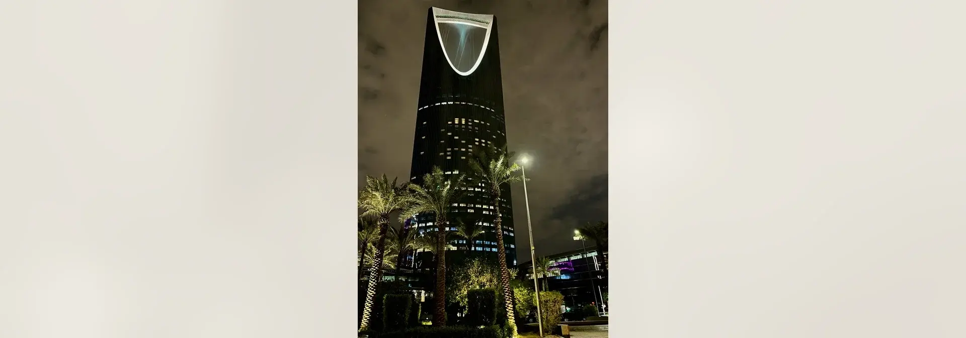 Kingdom Centre in Riyadh at night: the dark sloped tower with its illuminated inverted-arch crown rising above lit palm trees in the foreground garden