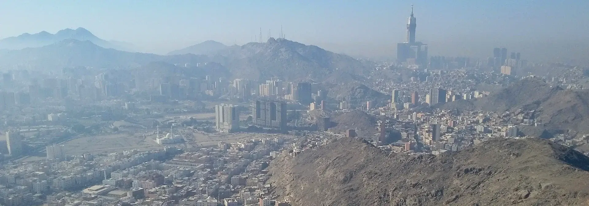 منظر جوي نهاري لمدينة مكة المكرمة يُظهر برج الساعة وأحياء المدينة الممتدة والجبال المحيطة