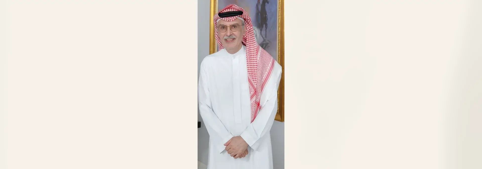 Prince Badr bin Abdulmohsen Al Saud wearing a red-and-white shemagh with a black agal, round glasses and a grey moustache, standing in a white thobe in front of a gold-framed painting