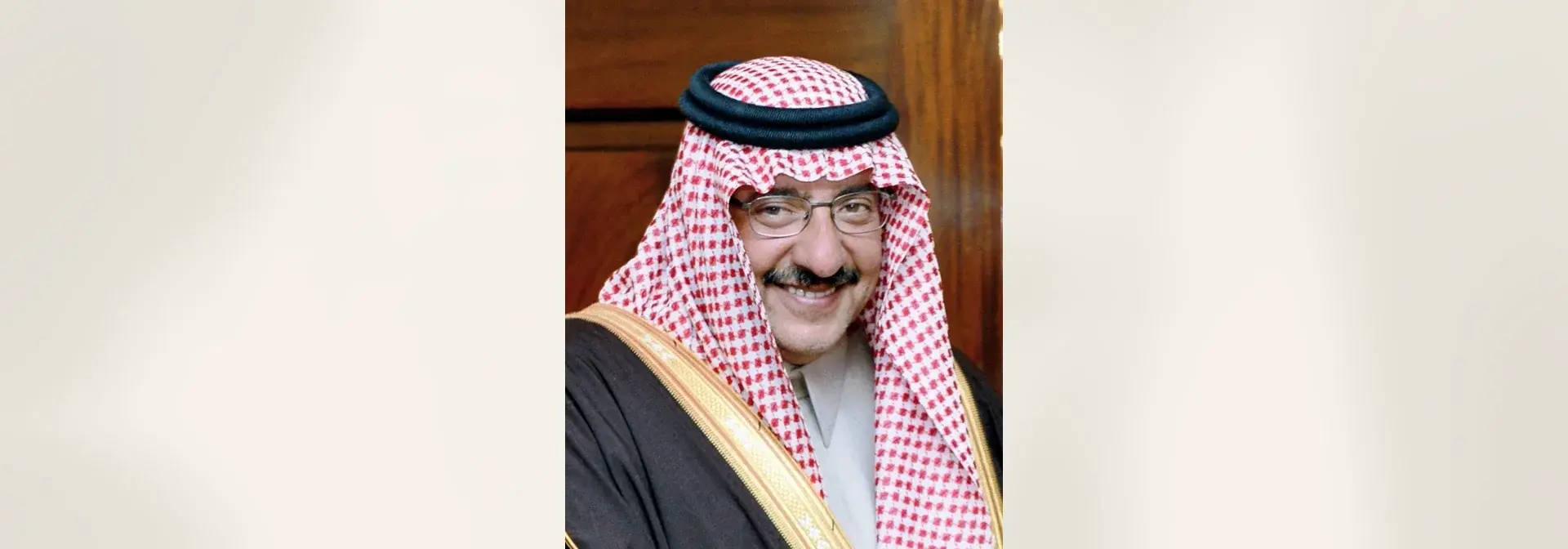 بورتريه الأمير محمد بن نايف بن عبدالعزيز بغترة حمراء وبيضاء وعقال أسود مزدوج ونظارة، مبتسماً بشارب كثيف ومرتدياً بشتاً أسود بحاشية ذهبية أمام باب خشبي في مبنى رسمي