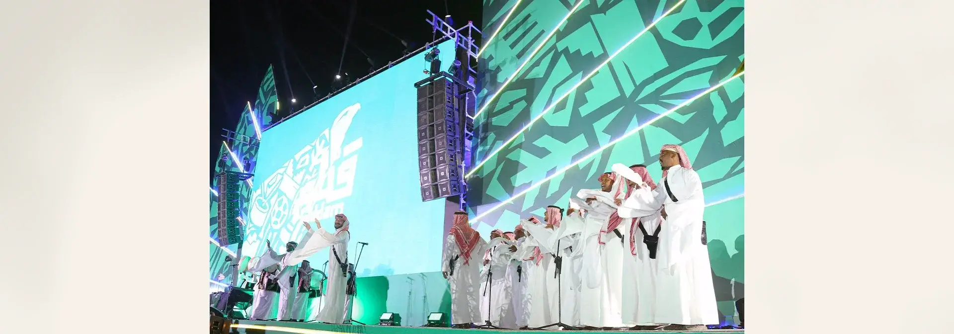 A live Samri folk performance on a lit stage at night: a line of Saudi men in white thobes, red-and-white shemagh, igal and bisht clap and sway, with two lead performers raising their arms in front
