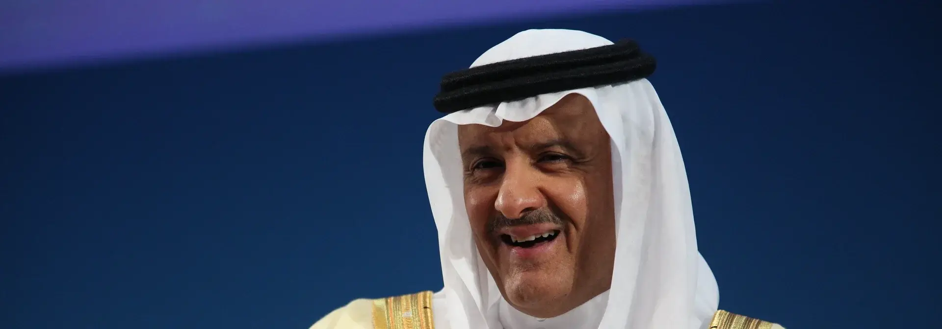Prince Sultan bin Salman bin Abdulaziz Al Saud smiling in a white ghutra and black double agal, the gold-trimmed collar of his cream bisht at the bottom of the frame, against a dark blue backdrop