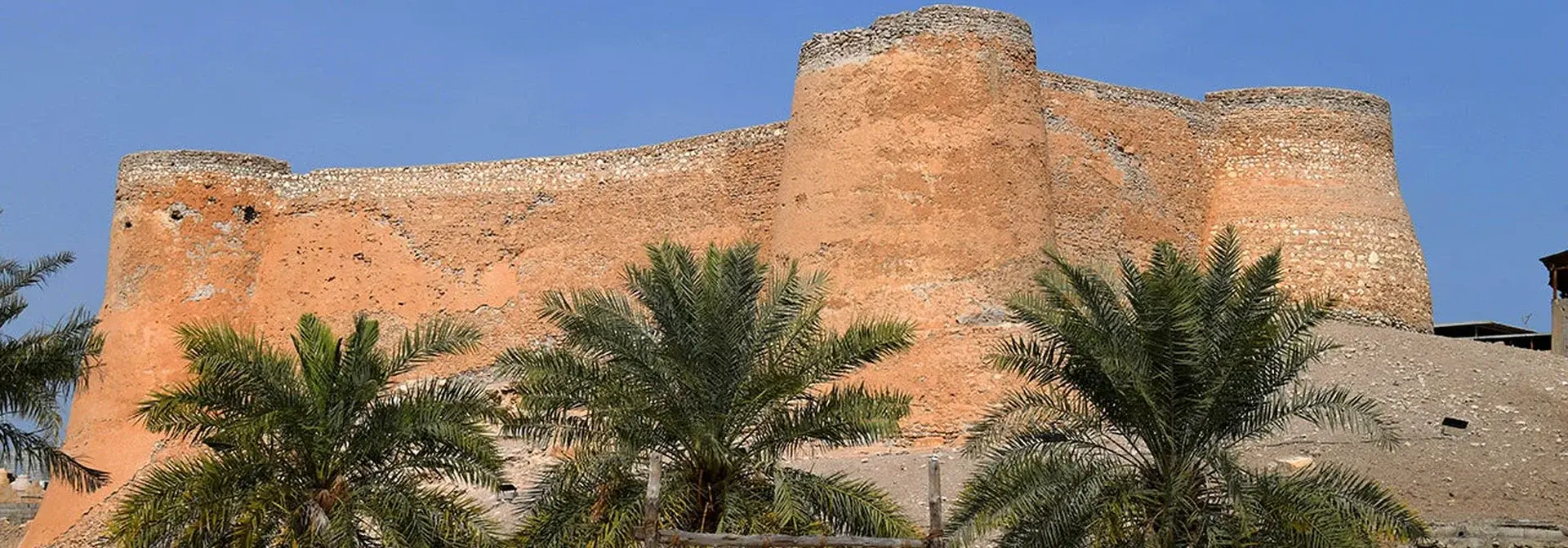 Tarout Castle on Tarout Island (Qatif, Saudi Arabia): the weathered orange mud-brick rampart with its round towers and three date palms in front, under a clear blue sky.