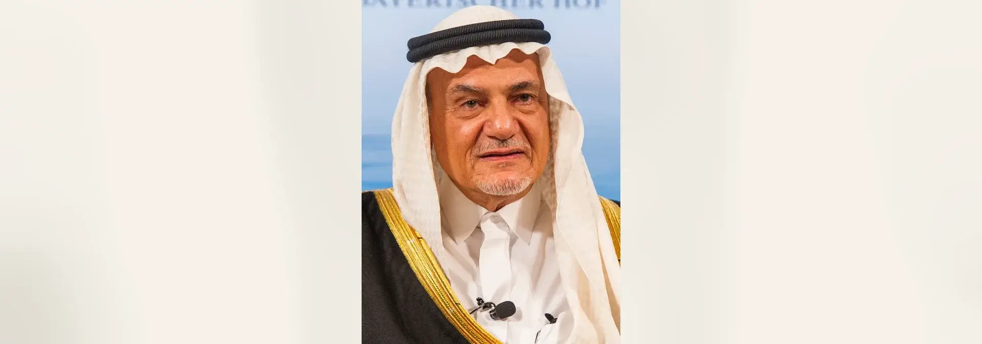 بورتريه الأمير تركي الفيصل بن عبد العزيز آل سعود، رجل بسحنة فاتحة ولحية خفيفة بيضاء يلبس غترة بيضاء وعقالاً أسود مزدوجاً وبشتاً أسود بحاشية مذهّبة فوق ثوب أبيض، يبتسم أمام خلفية زرقاء فاتحة