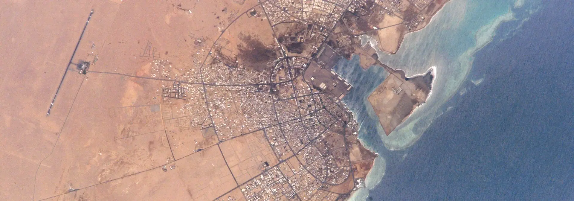 منظر جوّي لمدينة ينبع على ساحل البحر الأحمر يظهر النسيج العمراني الكثيف وشبكة الطرق والميناء ومهبط مطار ينبع والمرجان على حافة الساحل